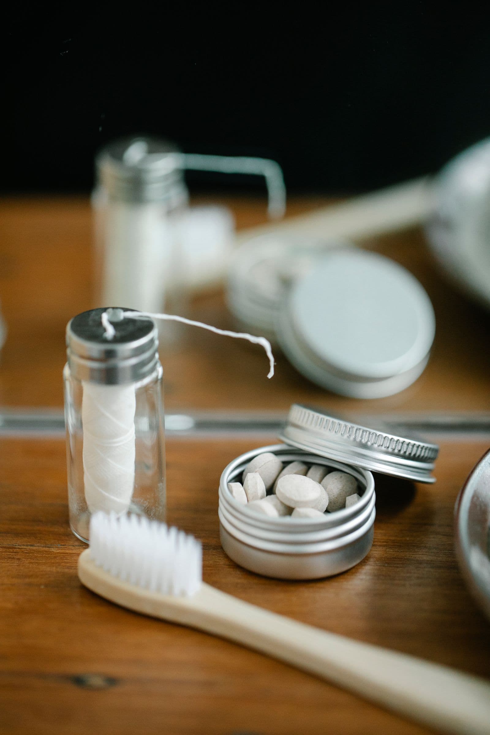 A bamboo toothbrush beside a glass jar of dental floss and a tin of toothpaste tablets on a wooden shelf